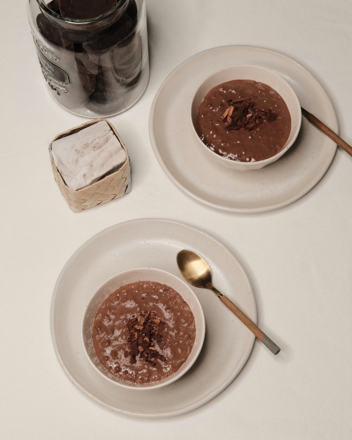 Two bowls of TSCS champorado with shaved chocolate beside a jar, spoons and a woven basket on a cream table.